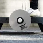 A person in black pants and white sneakers exercises on an Exxentric kBox Lite Starter System, featuring a central round pulley on a gray platform against a neutral background.