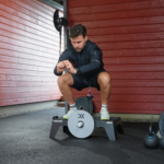 A man in athletic wear uses the Exxentric kBox Lite Starter System outdoors, adjusting his wrist strap. Nearby are a kettlebell and other workout gear, with a red wooden wall in the background.