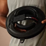 A person with a sleeveless shirt holds the Iron Neck 3.0, a black round fitness device featuring a strap with red and white stripes and a handle.