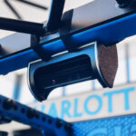 The Perch Camera, featuring a close-up of its lens, is mounted on a blue-painted metal frame with straps. The blurred background highlights the word CHARLOTT.