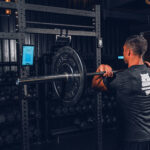 In a gym setting, a person wearing a dark t-shirt readies to lift a weighted barbell from the rack. In the background are kettlebells, weights, and a hanging rope. This is all captured by the Perch Camera as they face away from the lens.