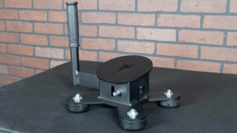 The Spring Ankle, a black three-legged device with a vertical handle, round base, leg bolts, and a slot on its main body, sits on a dark surface in front of a brick wall.