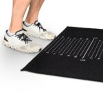 A person wearing white athletic shoes stands next to the OVR Roller Magnet (Copy), a black mat with white numbered lines, resembling a measurement or training tool on a white background.