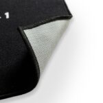 A close-up of the OVR Board Jump black floor mat shows its corner lifted, revealing a textured, non-slip gray backing and stitched black edges against a white background.