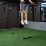 In a gym with mirrored and dark-painted walls, someone in shorts and athletic shoes leaps mid-air above artificial turf and two rectangular black-and-blue OVR Performance Jump blocks on the ground.
