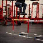 Wearing OVR Performance Jump athletic gear, a person leaps over a red and white hurdle in a gym, with weightlifting equipment and racks in the background.