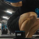 A person squats in a gym while holding a barbell, with the OVR Performance VBT device attached to the ground and connected by a cable to their lower body.