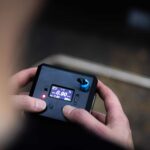 A person holds the OVR Performance VBT, a black device with a small display, buttons, knob, and red indicator light visible against a blurred background.
