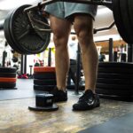Wearing black Nike sneakers, a person with muscular legs in shorts lifts a loaded barbell on a wooden platform at the gym, surrounded by weight plates and equipment, using the OVR Performance VBT for training.