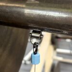 Close-up of the OVR Roller Magnet—a metal shackle connected to a thick bar, with a black swivel and small blue magnet hanging below. The blurred background shows gym or industrial equipment.