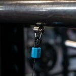 Close-up of the OVR Roller Magnet cable attachment on gym equipment, showing its metal bar, pulley connector, and a small blue magnetic piece secured to a cable. The blurred background hints at an indoor fitness environment.