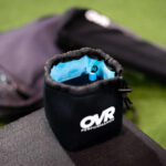 The OVR Velocity Case, a black drawstring pouch with OVR PERFORMANCE printed on it, rests on a surface, partially open to reveal its blue interior and a small blue item inside; in the blurred background are a bag and green turf.