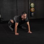 A man performs a bodyweight exercise on a flat bench, using the VALD NordBoard placed on a black rubber mat, with colorful weight plates visible on the wall behind him.