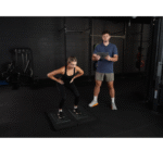 A woman performs a squat on exercise mats in a gym while a man with a clipboard observes her using VALD ForceDecks, with various gym equipment visible in the background.