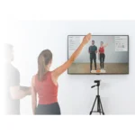 A woman raises her right arm while following a workout on a wall-mounted TV featuring two instructors. A man stands beside her, and a VALD HumanTrak camera system on a tripod faces them.