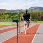 A person uses the OVR Performance Sprint Starter Kit on an outdoor track with timing sensors, surrounded by hills and greenery beneath a partly cloudy sky.