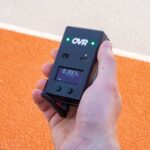 A hand holds the OVR Performance Sprint Starter Kit—a black device with OVR branding, small display, USB-C port, buttons, antenna, and two green lights—set against an orange and beige textured background.