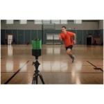 A man in an orange shirt and black shorts is running indoors on a gymnasium court, with a motion sensor device on a tripod recording his movement in the foreground.
