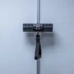 The Ancore Mounts, featuring a strap handle, is secured to a vertical metal pole against a smooth light-gray wall. This black exercise resistance attachment has measurement markings and a sturdy, compact design.