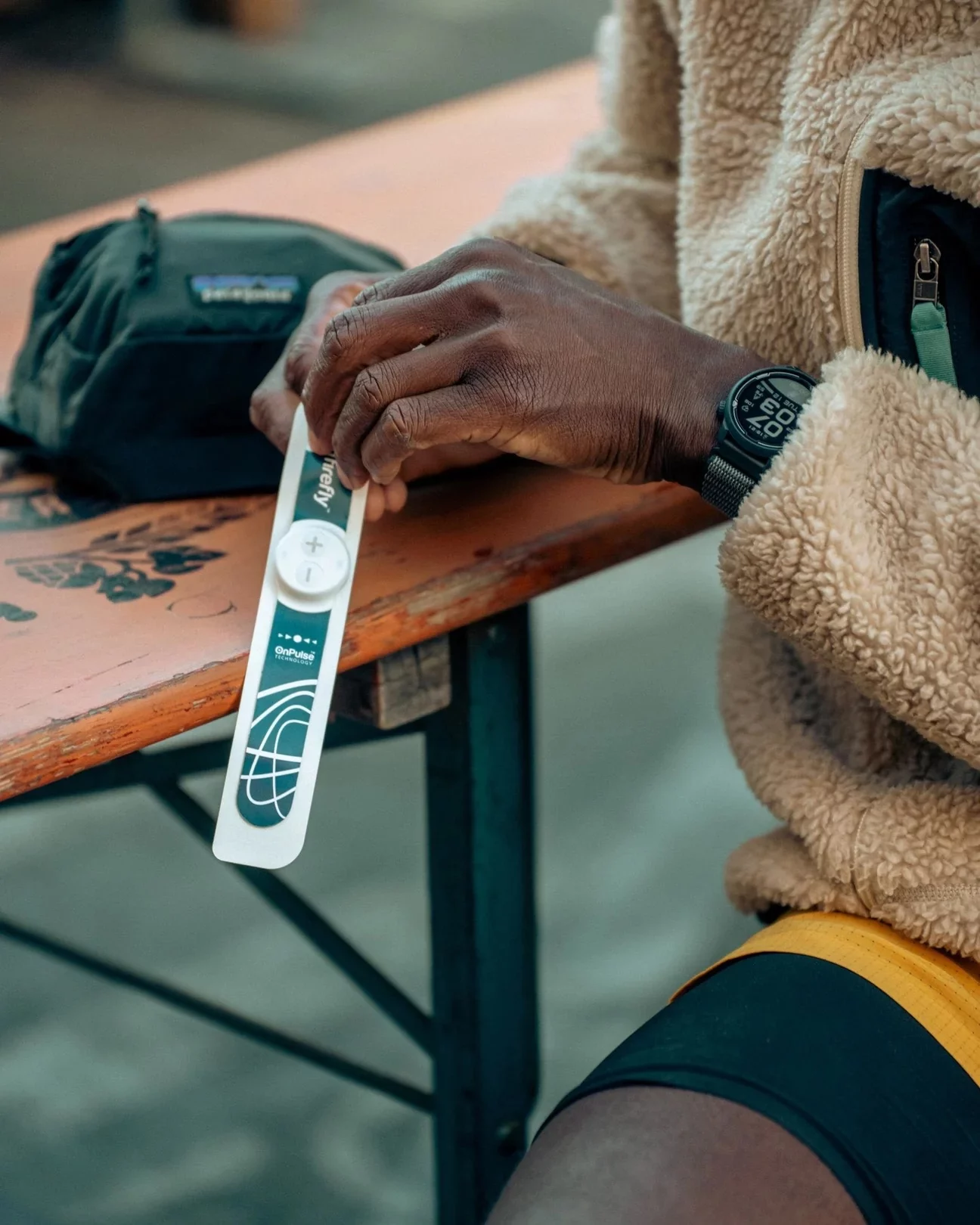 A person wearing a tan fleece jacket and watch sits at a wooden table, holding a white and green COVID-19 rapid test device. A small black bag rests on the table.