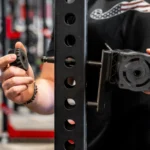 Wearing a black shirt with a flag design, a person attaches an Ancore Mount to a black metal rack with evenly spaced holes, aligning its round component with the threaded rod for secure installation.