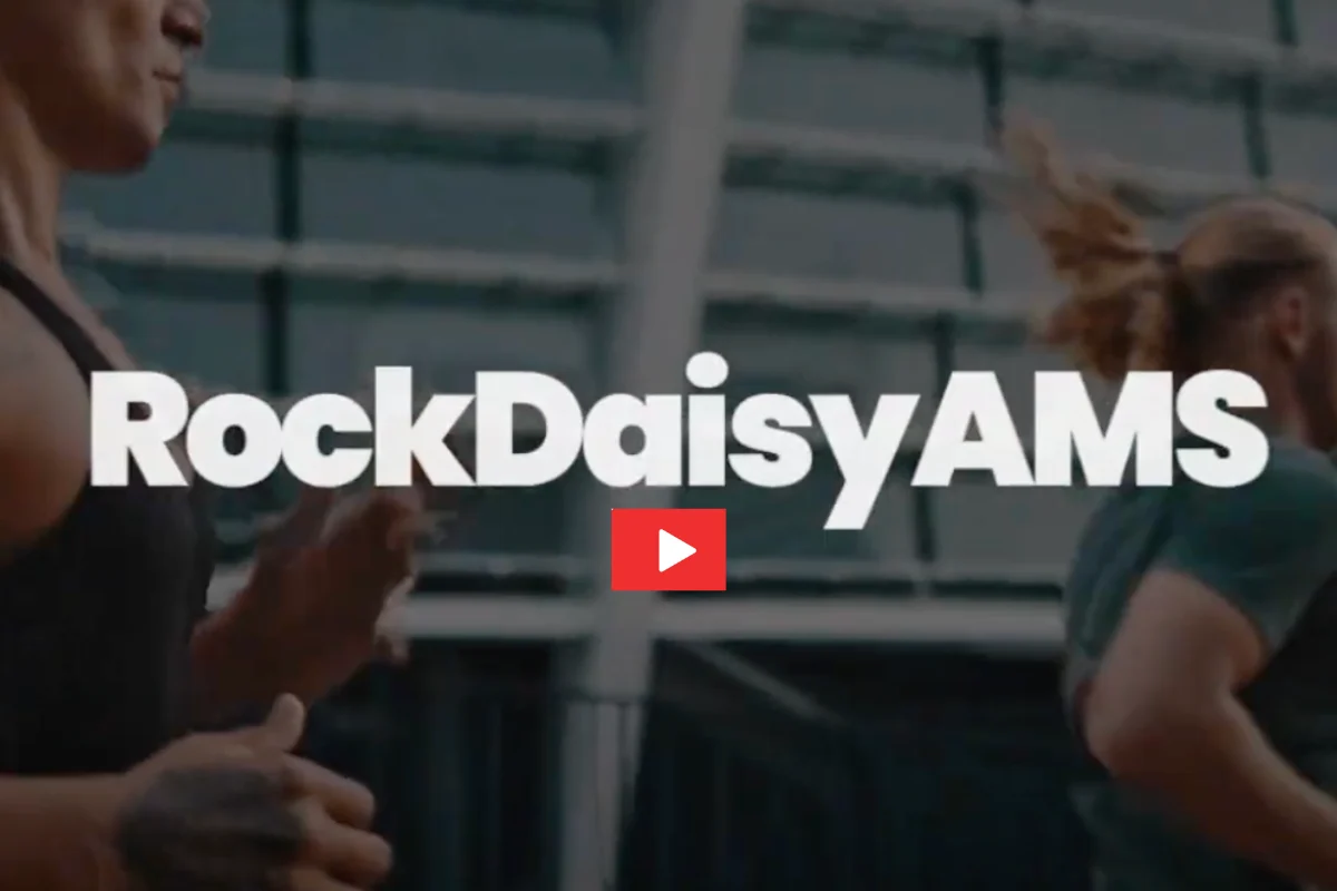 Two people run outdoors in front of a blurred building. RockDaisy AMS appears in bold white letters at the center, with a red play button icon below the text.