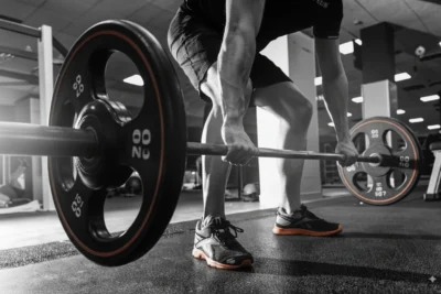 A person in athletic shoes and shorts grips a barbell loaded with weights, preparing to deadlift in a gym. The image is mostly black and white, highlighting the barbell and shoes in color.