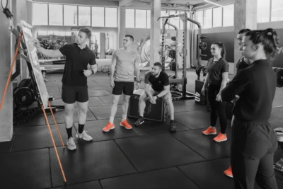 A fitness instructor explains something on a whiteboard to five gym-goers, all wearing black outfits with bright orange shoes, in a gym with various workout equipment.