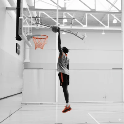 A basketball player in mid-air dunks the ball into an indoor hoop. The image is mostly black and white, with the players shoes, shorts, and the basketball rim highlighted in orange.