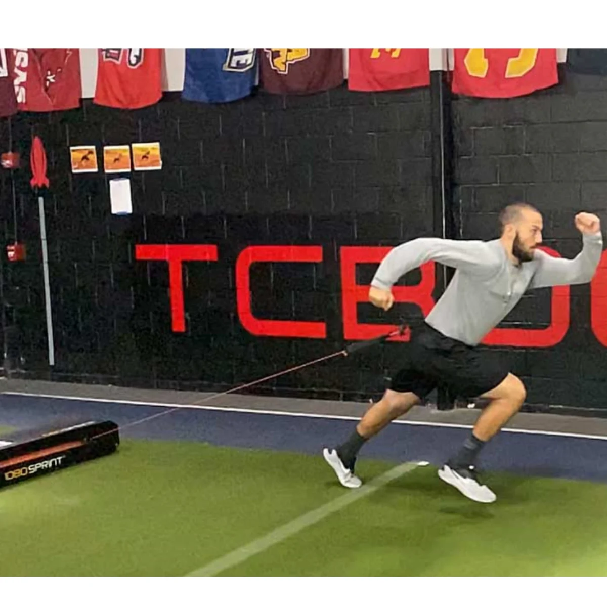 A man in athletic wear sprints forward on turf while attached to a resistance band at a training facility. Sports team banners hang on a black wall behind him with large red letters.