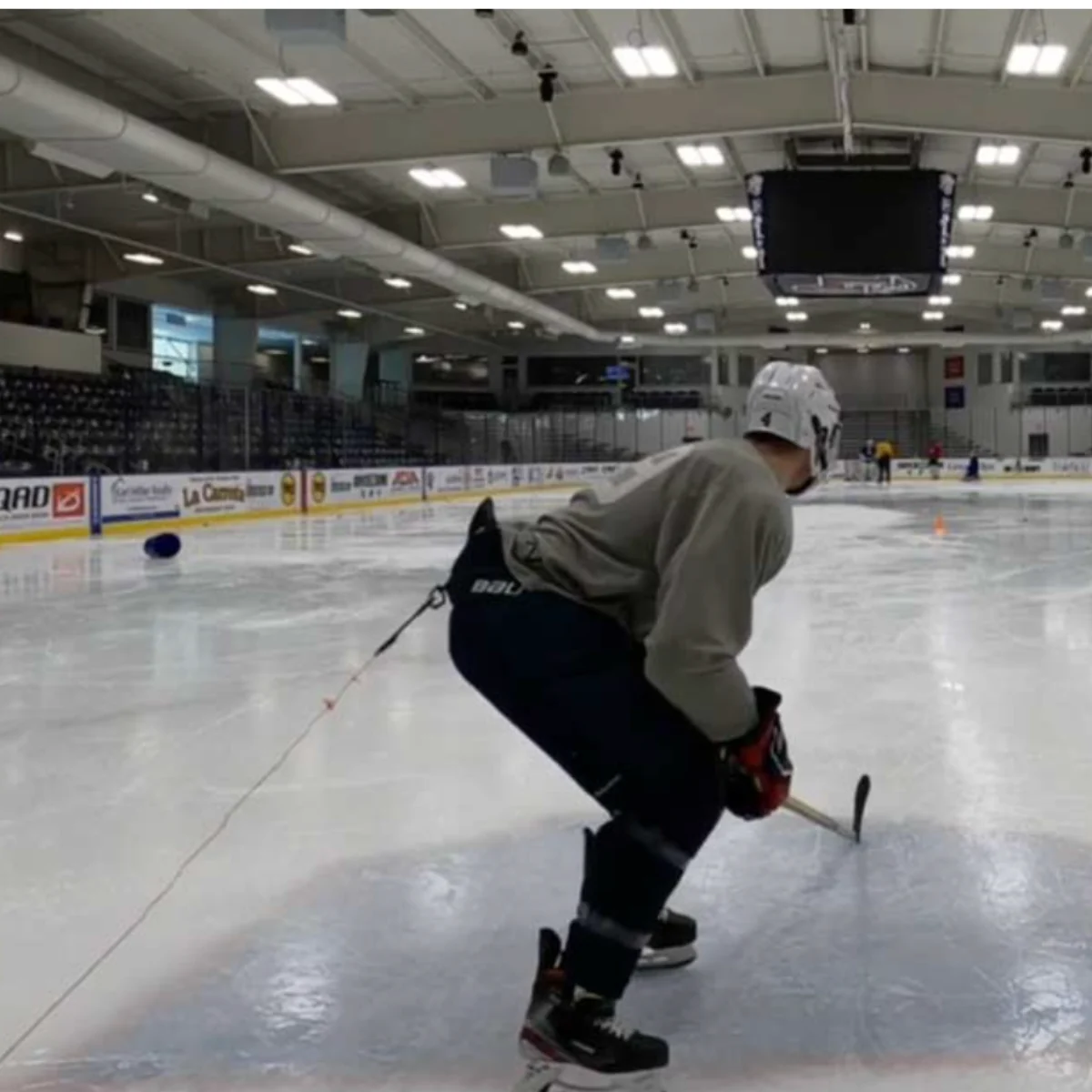 A hockey player in full gear skates on an indoor rink, bending forward with a stick and puck, attached to a resistance cord. The arena is well-lit with empty seats and a scoreboard overhead.