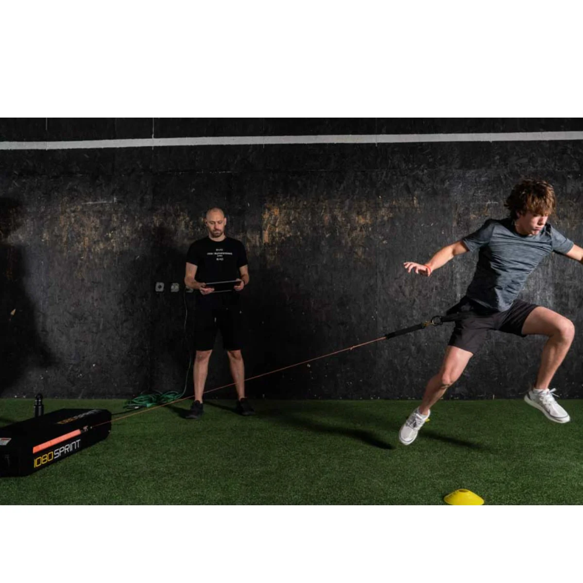 A young man wearing athletic clothes leaps forward with a resistance band attached to his waist, while another man stands in the background holding a device, observing him on a turf surface indoors.