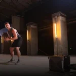 Wearing TAPEX athletic gear, a man trains indoors with resistance bands attached to his waist and anchored to a heavy box. The scene features large concrete columns and warm lighting in the background.