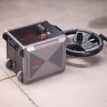 A gray, box-shaped TAPEX machine sits on a concrete floor beside two stacked black weight plates. It features wheels, control buttons, and a red emergency stop button on top.