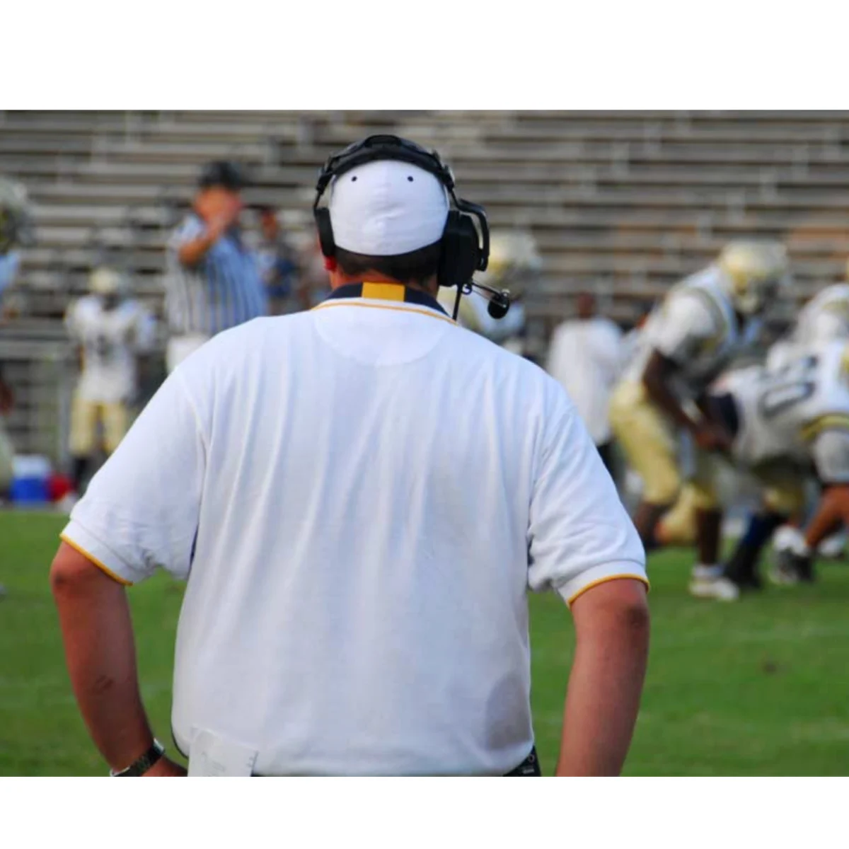 A football coach wearing a headset and white shirt stands on the sideline watching a game, with players and a referee visible in the blurred background on the field.