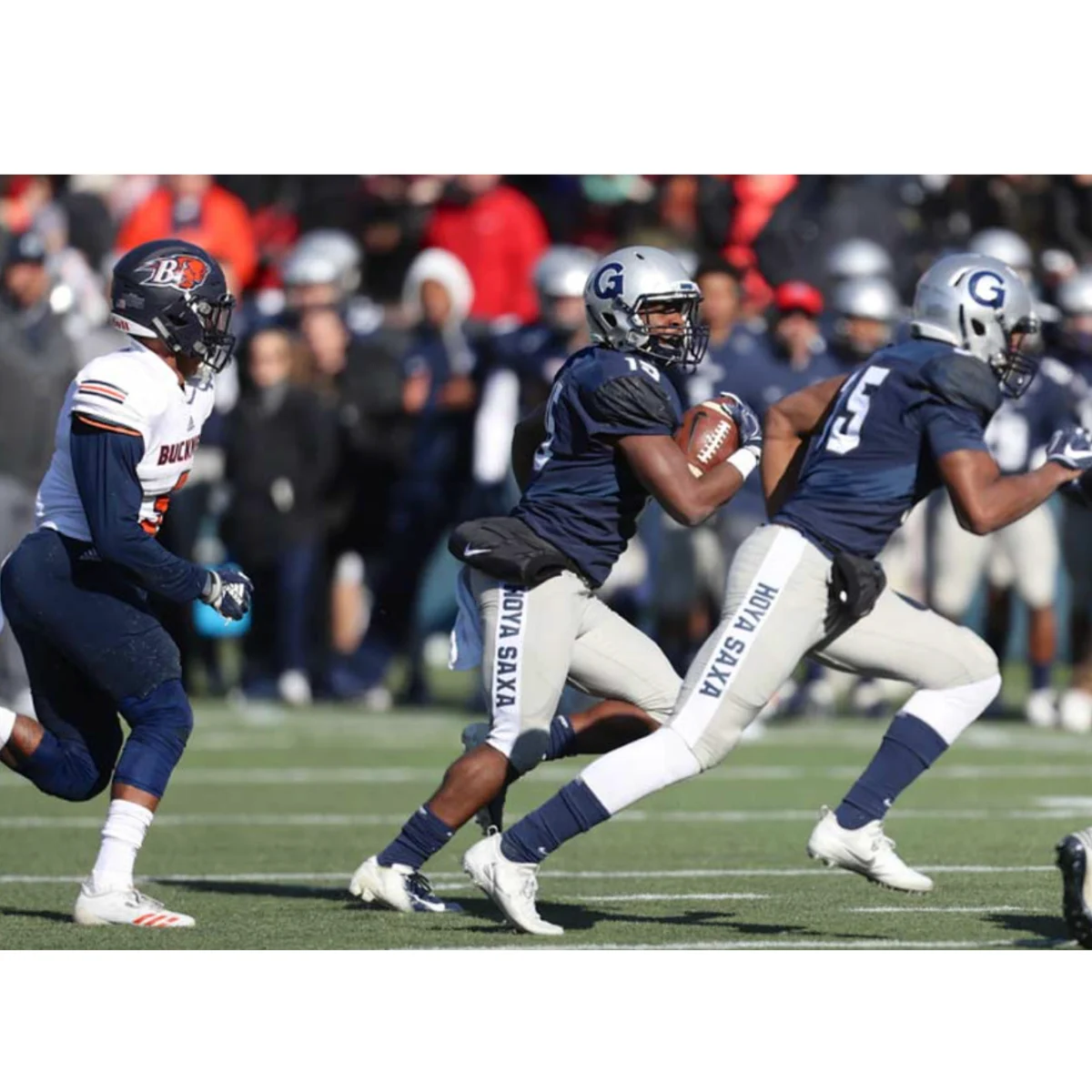 A football player in a blue and gray uniform runs with the ball, followed by teammates, while an opposing player in a white jersey chases them on the field. Spectators are visible in the blurred background.