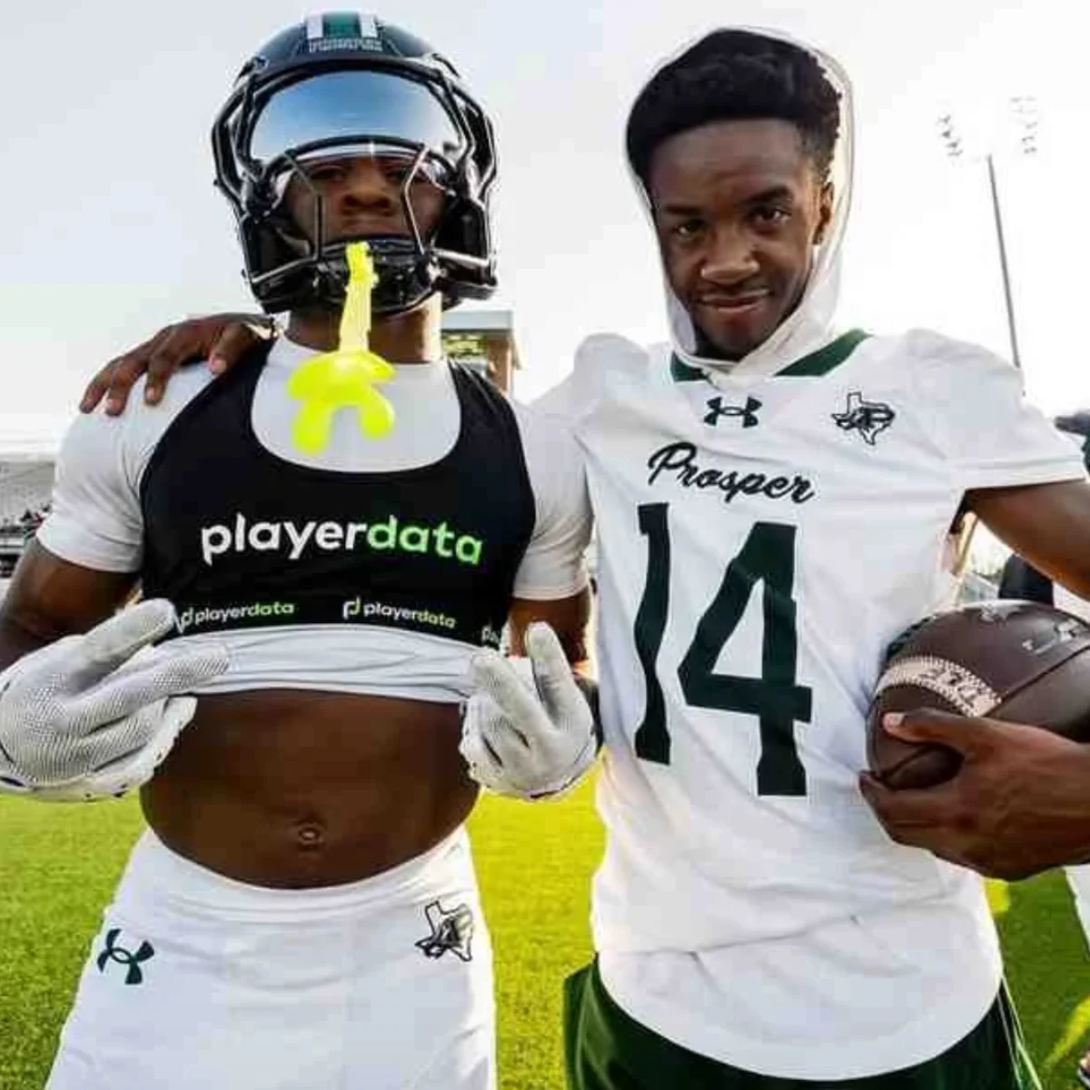 Two American football players in white uniforms pose on a field. One lifts his shirt to show a chest monitor, while the other, holding a football, stands beside him smiling. They appear relaxed and confident.