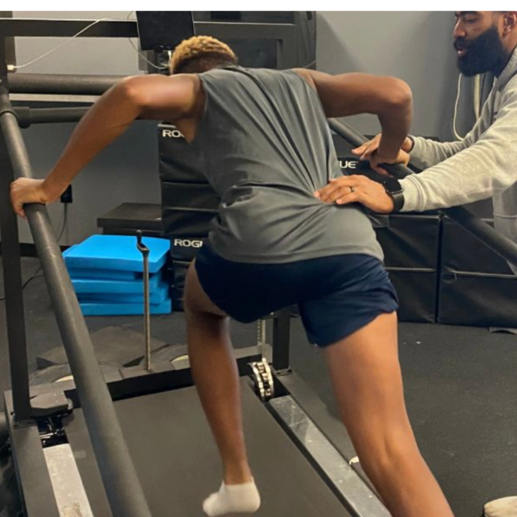 A person in athletic wear runs on a treadmill at an incline, gripping the handrails, while another person stands next to them, offering support and guidance. Blue foam pads and gym equipment are visible in the background.