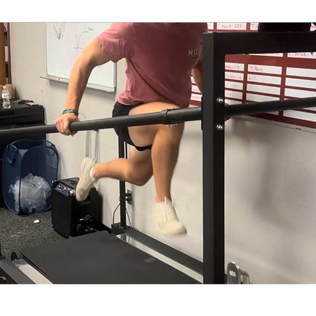 A person in a pink shirt and white shoes is mid-jump over a horizontal metal bar in a gym or fitness room, with a whiteboard and a trash bin in the background.