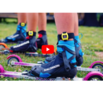 Close-up of people ready for roller skiing on grass, wearing blue and black boots, with the Freelap Biatholon Strap visible. A red play button is overlaid in the center of the image.