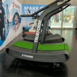 Three TRUEFORM.TRAINER™ Curved Treadmill Turf units with green running surfaces are lined up indoors, and a banner featuring runners is visible in the background.