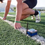 A runner crouches at the starting line on a sports field, wearing athletic gear and colorful shoes. A small electronic timing device with an antenna is placed on the ground beside the runner.