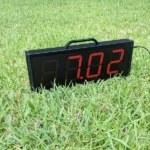 The Dashr Display is set on a grassy field, showing 7.02 in red. It features a black handle on top and has a visible power cord extending to the right.