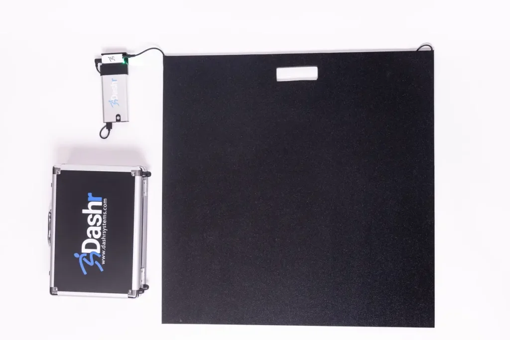 The Dashr Jump Mat and Controller, a black rectangular mat with an attached small electronic device, is shown next to a silver Dashr carrying case on a white background.