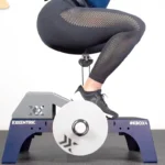 A person in black leggings and shoes squats on a kBox4 exercise device with EXXENTRIC and #KBOX4 markings. Their feet are on the platform, hands not visible. The scene includes the Exxentric Decline Board.