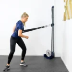 A woman in a gym uses the Exxentric Rotational Sling, performing resistance cable exercises. Shes wearing a blue shirt and black leggings, leaning slightly forward. The room features white walls and a carpeted floor.
