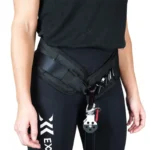 Wearing a black Exxentric Squat Hip Belt with metal carabiner and pulley system, a person in black leggings featuring a small logo on the thigh stands against a plain background.