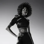 A woman with curly hair confidently poses for the camera in a sports bra and leggings, wearing a Hyperice Ice ICT Left Shoulder brace. The image is in black and white.