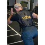 A person with gray hair, seen from behind, wears the OHM Power Harness black vest while sprinting indoors on a track with white lines; gym equipment appears in the background.