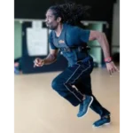 A man with long dreadlocks in a dark athletic outfit and sneakers runs indoors wearing the OHM Power Harness, appearing focused and in motion against a blurred background.